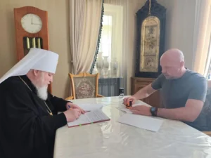 Два человека за столом: один в церковной одежде, другой в повседневной, обсуждают документы.